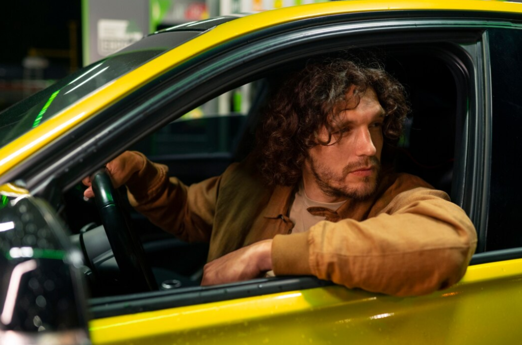 man with wavy dark hair looks outside of the yellow car, one hand on the car wheel, another  the second looks from the car