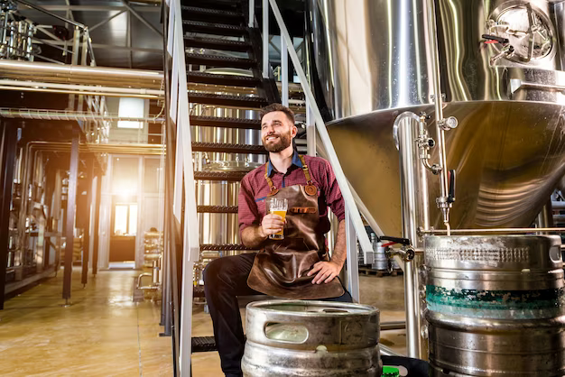 A man sits in a workshop among barrels and drinks beer