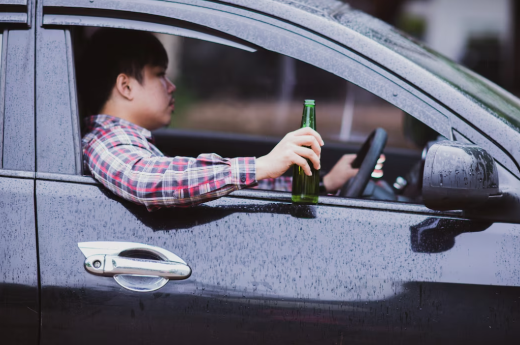 man in a colorful shirt sits at the car wheel and holds a bottle outside of the car, raindrops on the black car 